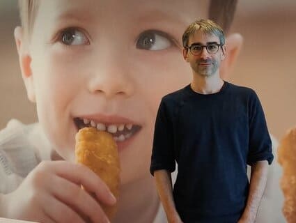A man stands in front of a large picture of a boy eating a chicken nugget. 一个男人站在一张男孩吃鸡块的大照片前。